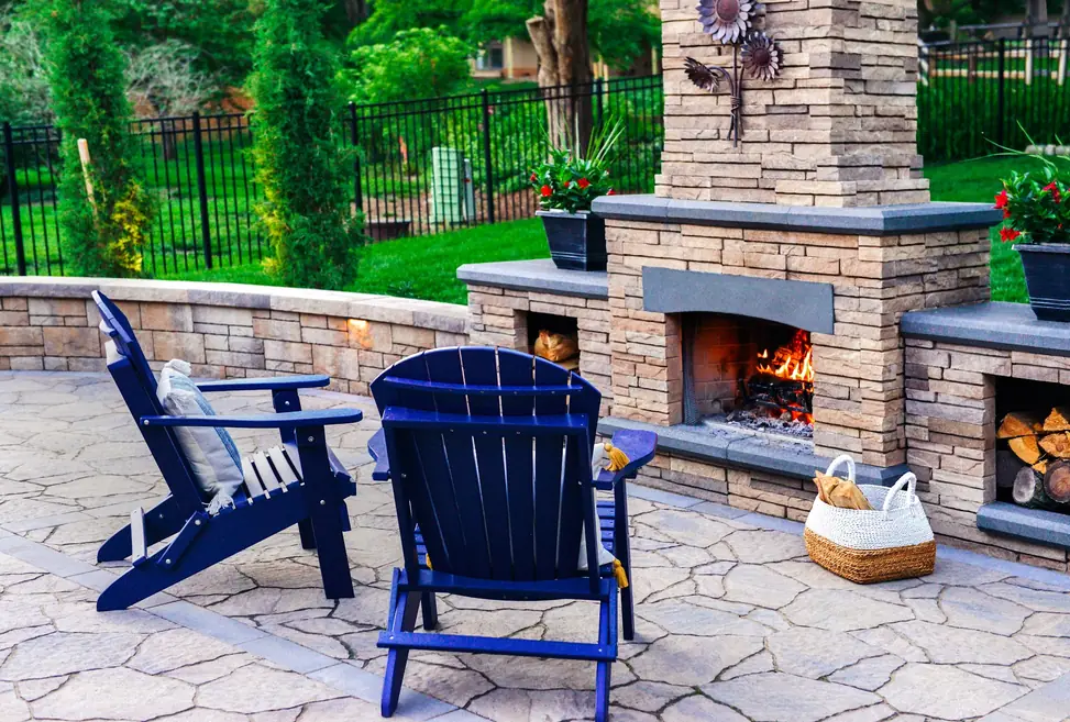 Stacked stone outdoor fireplace with a live fire burning, bluestone caps and navy Adirondack chairs on a flagstone patio